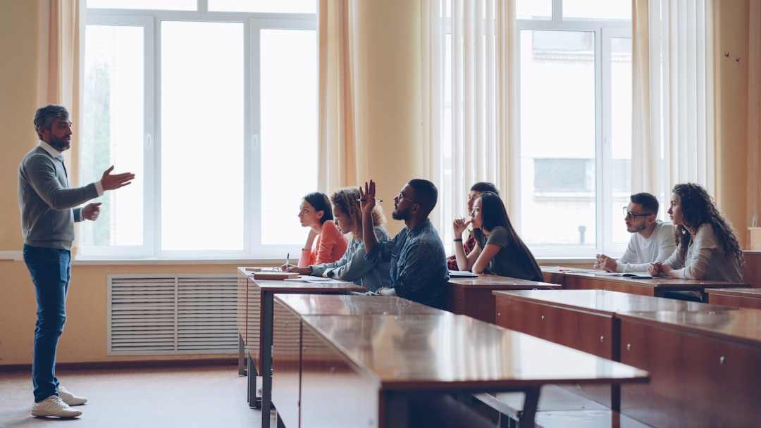 Friendly teacher is talking to students, smart man is raising hand and speaking sitting at table in spacious lecture hall while his classmates are writing.